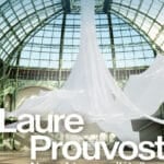 Poster for Laure Prouvost installation at Grand Palais, showing flowing white fabric sculptures in a glass atrium with the artist's name and exhibit title text (Nous, frissons d'étoiles).
