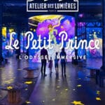 Interior view at Atelier des Lumières advertising Le Petit Prince: immersive exhibit with purple-blue projection, starry floor, and visitors standing before a large Celestial display.