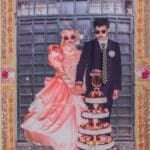 A couple dressed formally, woman in a pink gown and sunglasses and man in a dark suit with a striped tie, standing beside a multi-tier cake outside a gate.