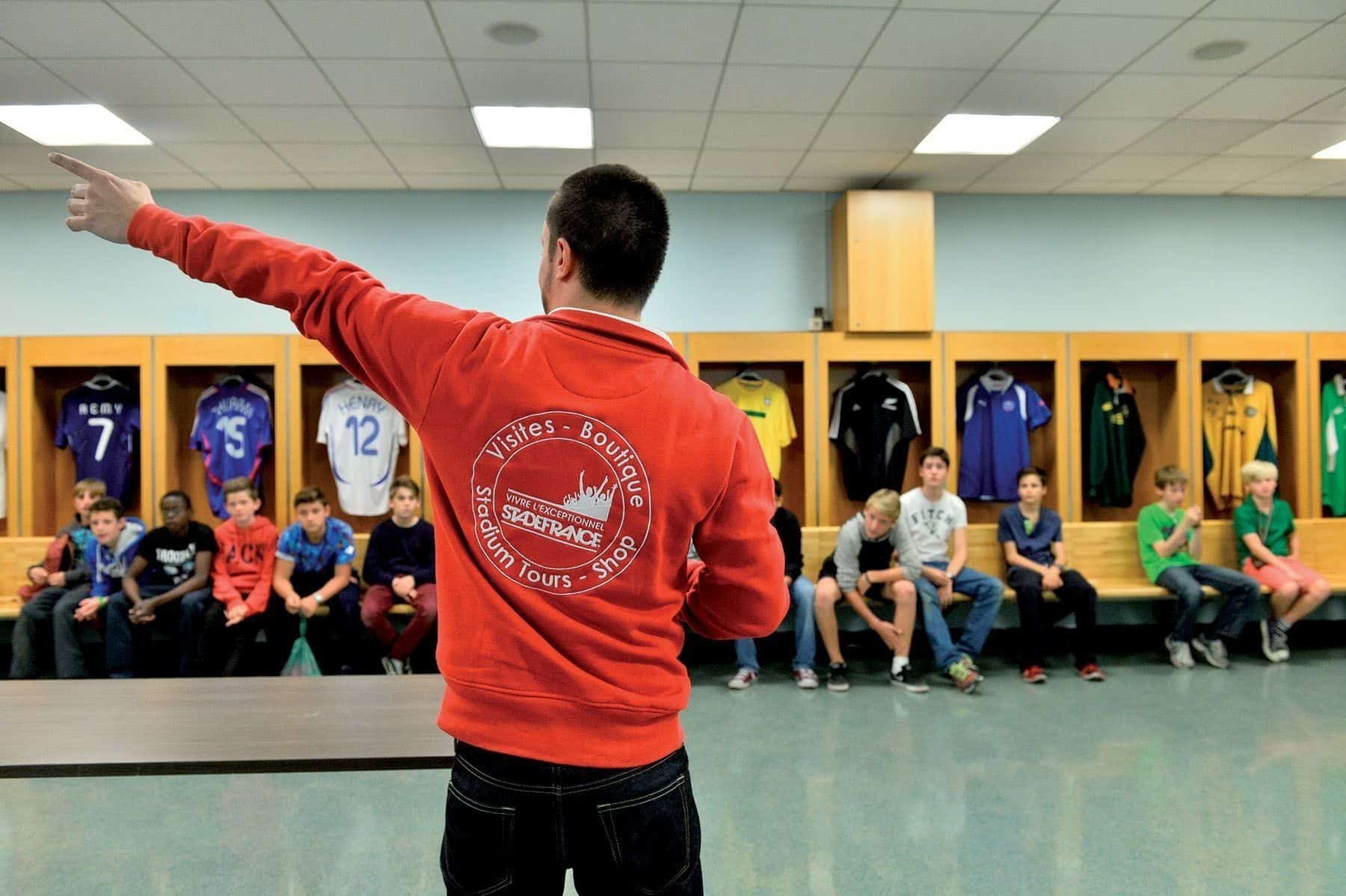 Guide et groupe de visiteurs dans un vestiaire des joueurs, maillots suspendus aux casiers